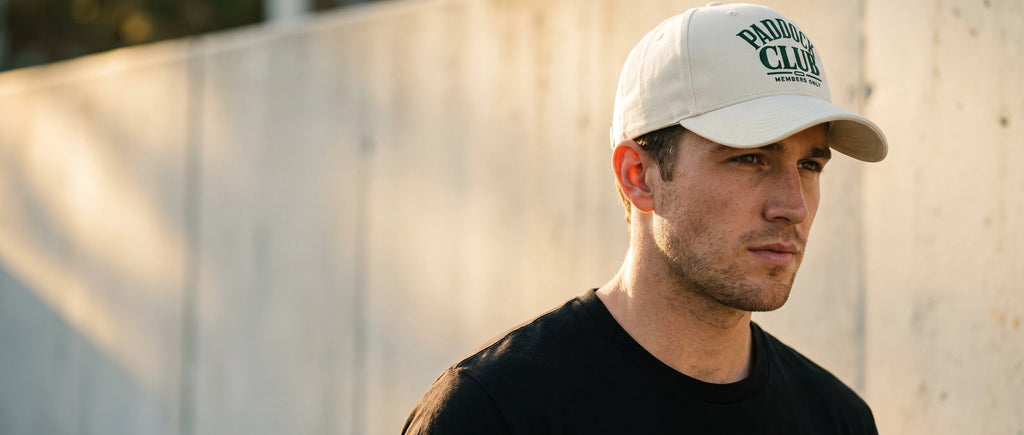 Homme portant la casquette Paddock Club crème devant un mur de béton au soleil couchant