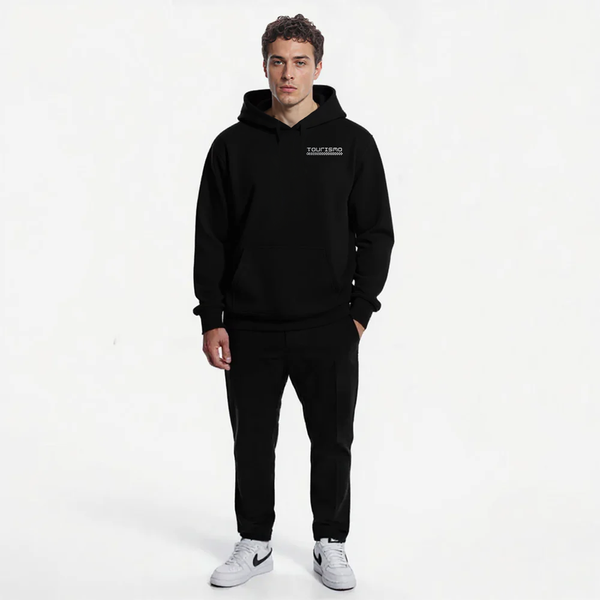 Sweat à capuche noir homme avec logo blanc Tourismo sur le cœur, pantalon noir assorti, baskets Nike blanches. Look urbain sobre. Modèle debout, une main dans la poche, fond blanc épuré.
