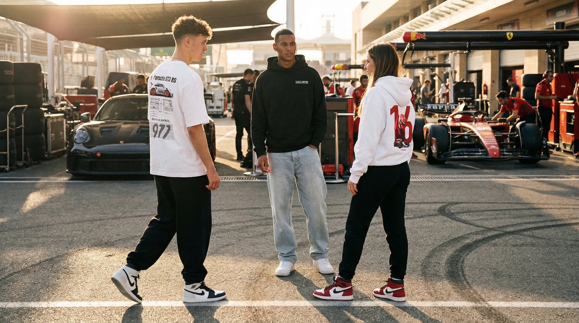 Trois mannequins Tourismo dans un paddock de course au coucher du soleil, portant un t-shirt Porsche GT3 RS, un hoodie noir Tourismo et un hoodie Ferrari Charles Leclerc, avec une Porsche et une Formule 1 Ferrari en arrière-plan.