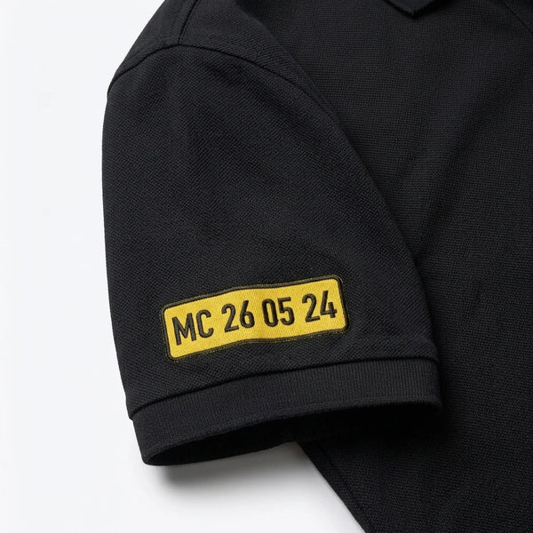 Gros plan sur la plaque MC 26 05 24 brodée sur la manche du polo noir